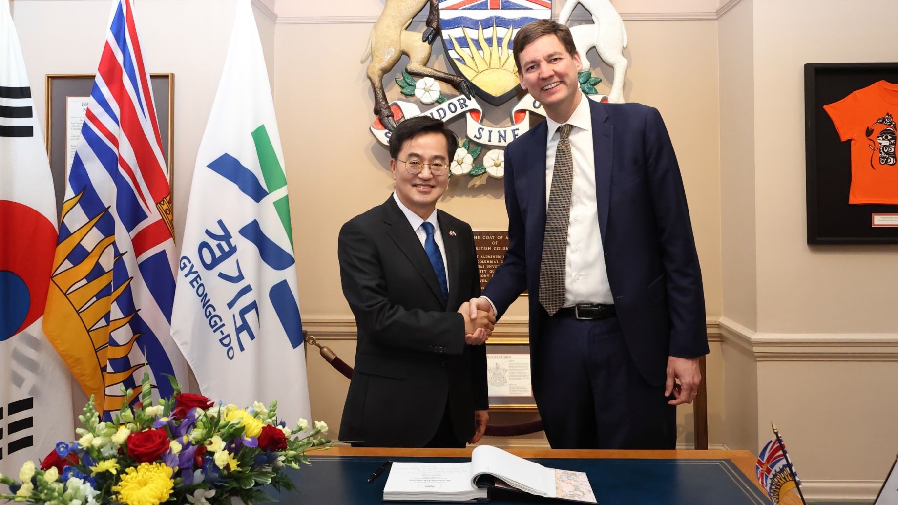 Premier Eby and Governor Kim shaking hands by a desk and flags. 